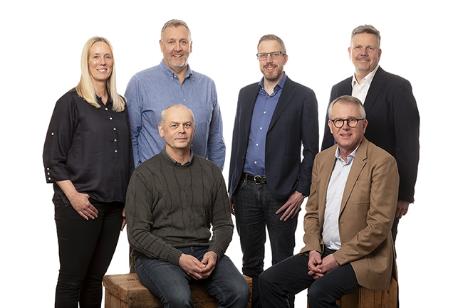 Gruppfoto av sex personer, tre stående och tre sittande, mot en vit bakgrund. Fyra män och två kvinnor är formellt klädda, med neutrala uttryck och avslappnad hållning.